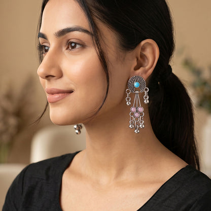 A woman models one oxidised silver long drop earring with pink and turquoise stones and ghungroo bells, looking left.