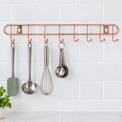 A rose gold metal wall hanger with eight hooks displays kitchen utensils against a white tiled background.