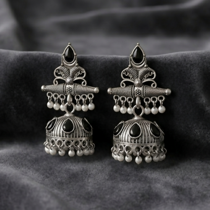 A pair of oxidized silver jhumka earrings with black stones and white pearls rests on a dark grey velvet surface.