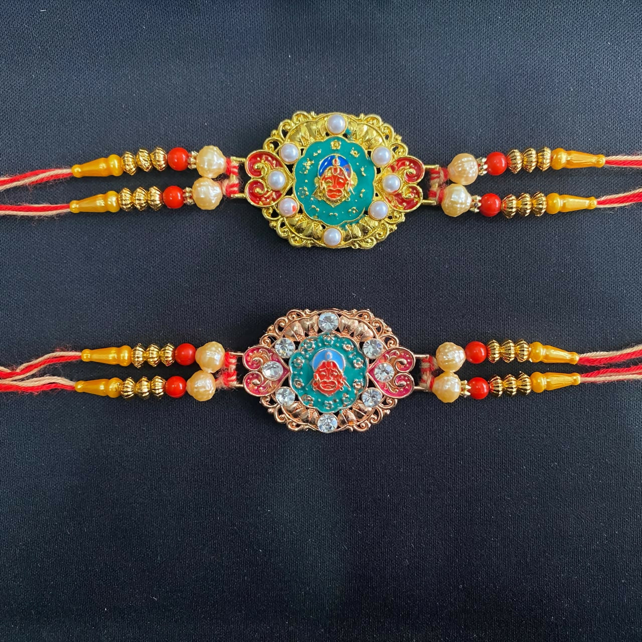 Two Hanuman Ji designer rakhis with gold and rose gold accents, pearls, and red threads, displayed on a plain black