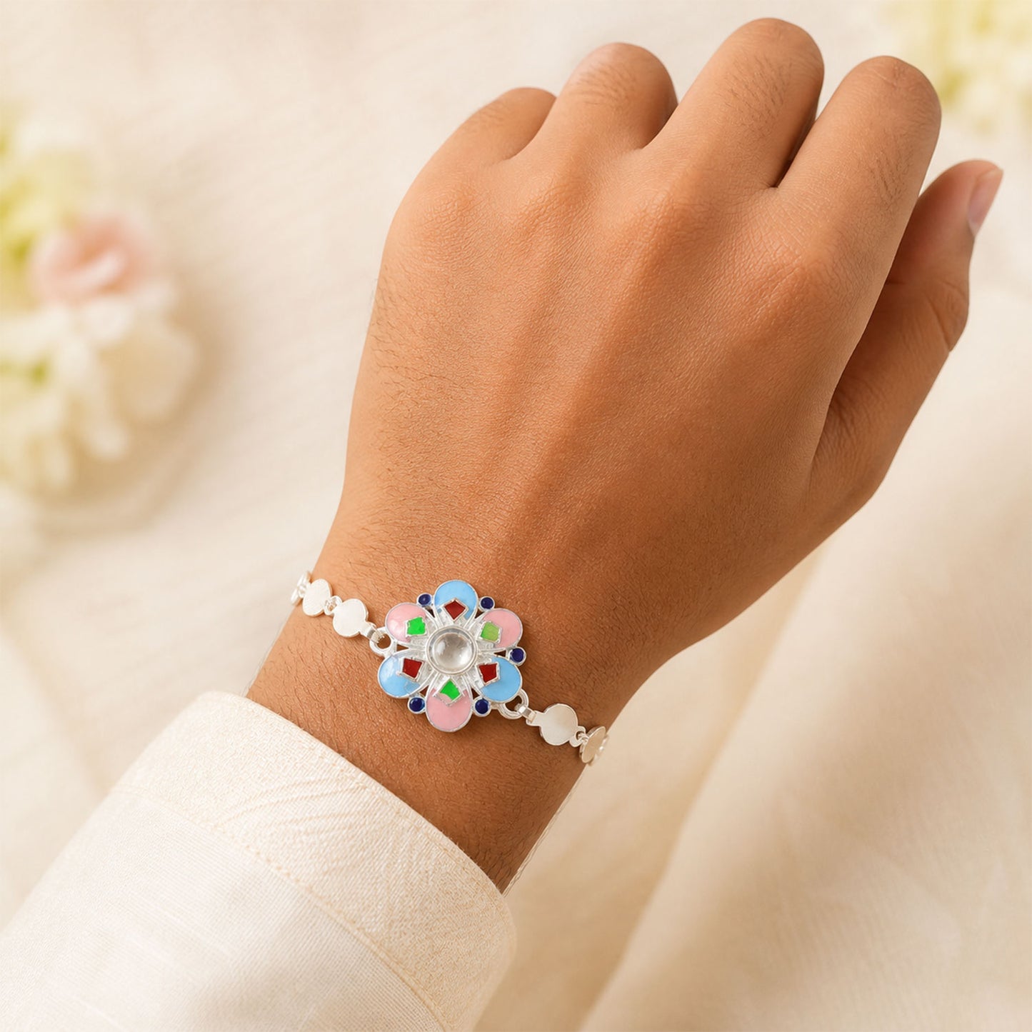 A man's wrist wearing a silver chain rakhi with a colorful enamel flower charm, against a white fabric.