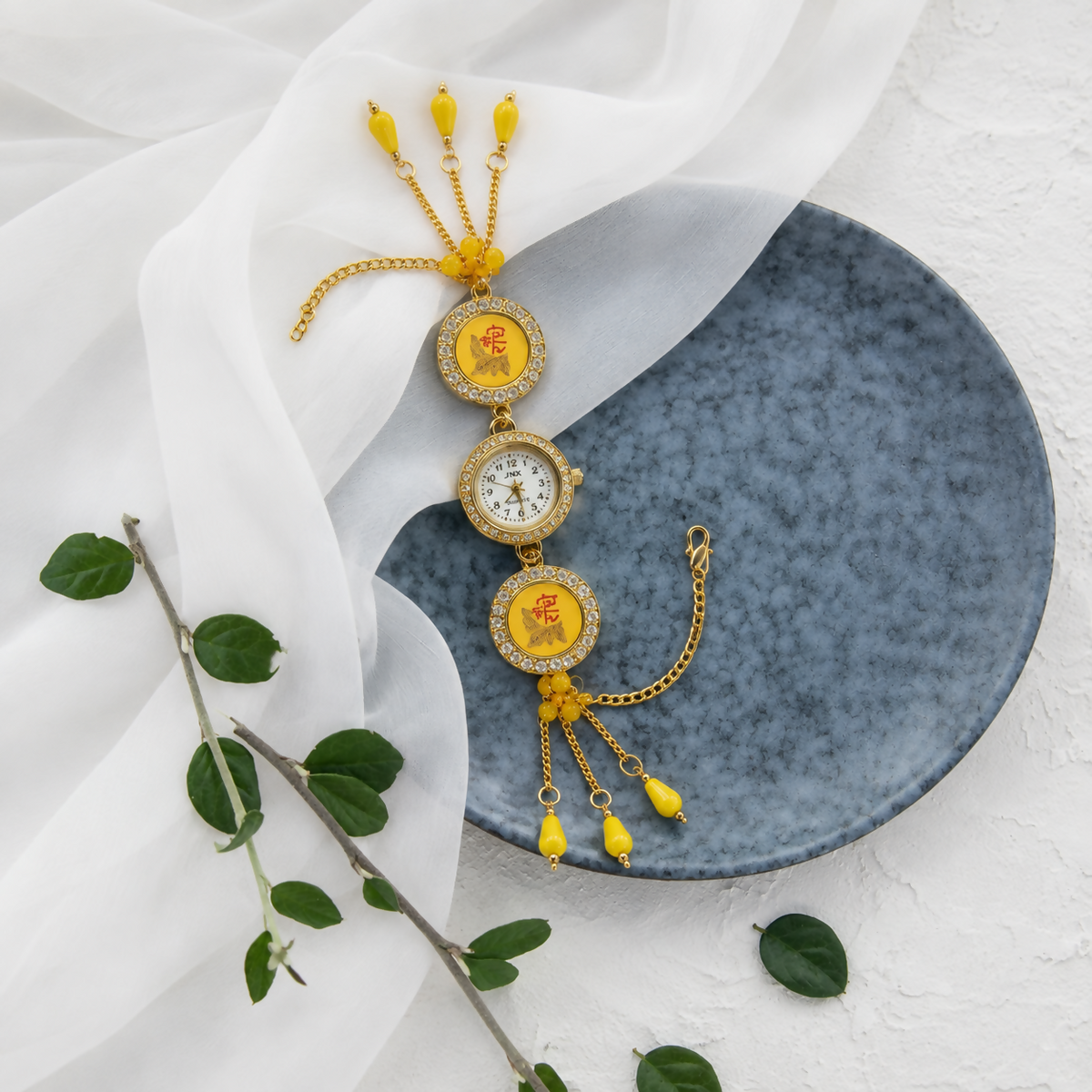 Gold-toned bracelet watch with yellow beads and charms, resting on a blue plate with a white sheer fabric and green leaves.
