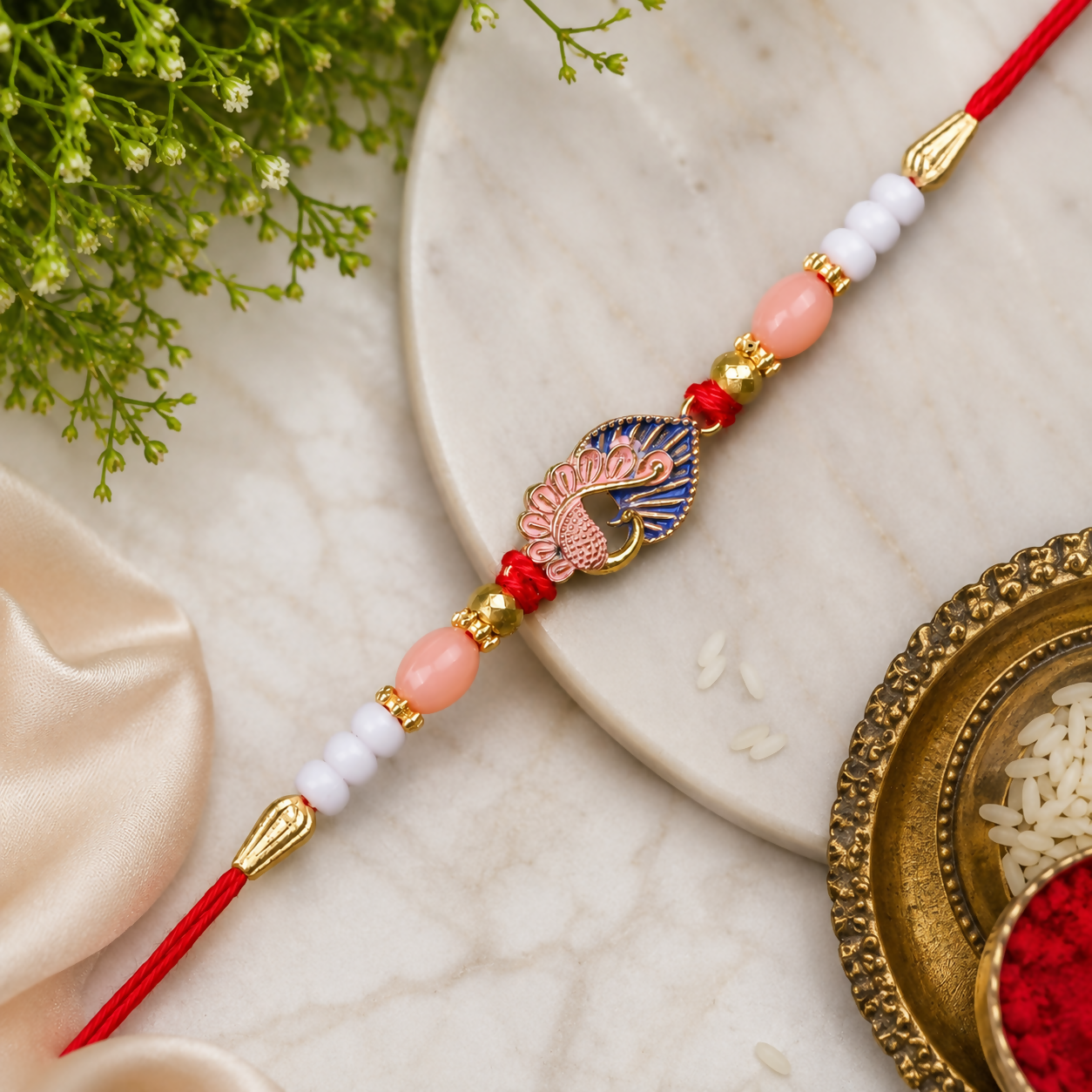 A red thread with a gold and blue peacock charm, flanked by pink, white, and gold beads, rests on a marble surface.