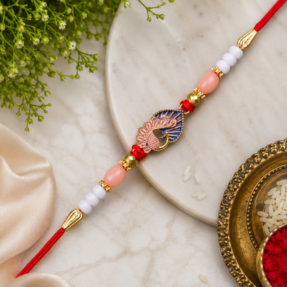 A red thread with a gold and blue peacock charm, flanked by pink, white, and gold beads, rests on a marble surface.