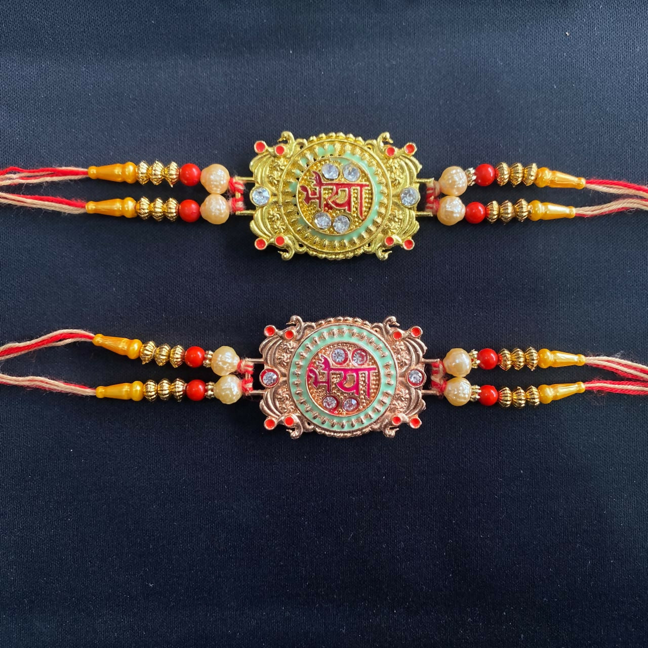 Two ornate Rakhis on a plain black background, one gold with green enamel, the other rose gold with pink, featuring red and
