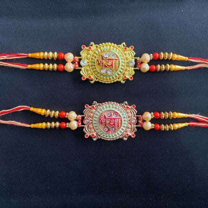 Two ornate Rakhis on a plain black background, one gold with green enamel, the other rose gold with pink, featuring red and