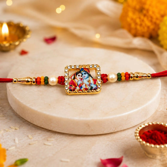 A gold-toned square Radha Krishna enamel charm with a crystal border and pearl accents is centered on a red thread with