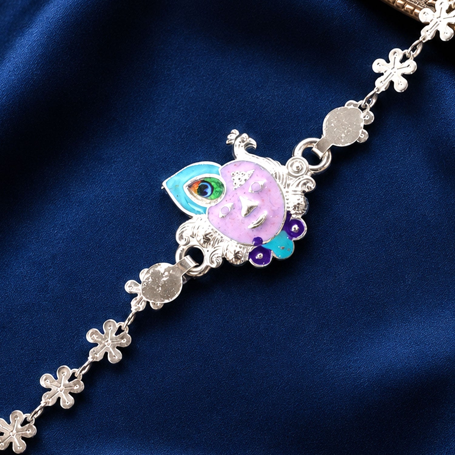 Silver rakhi featuring a purple enamel Krishna face and peacock feather charm against a deep blue satin fabric.