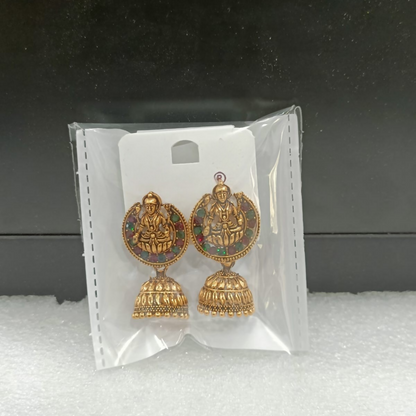 Two gold-toned jhumka earrings with goddess figures and colorful stones are packaged in a clear plastic bag.