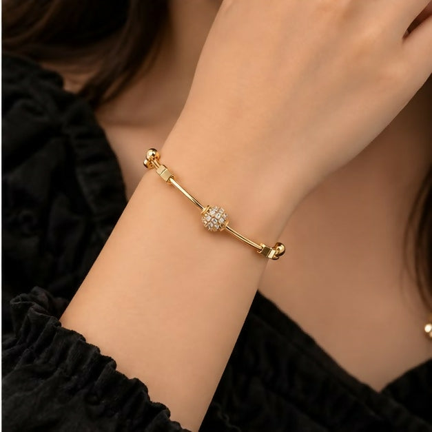A woman's wrist adorned with a gold-tone adjustable bangle featuring a dazzling crystal ball charm, against a dark top.