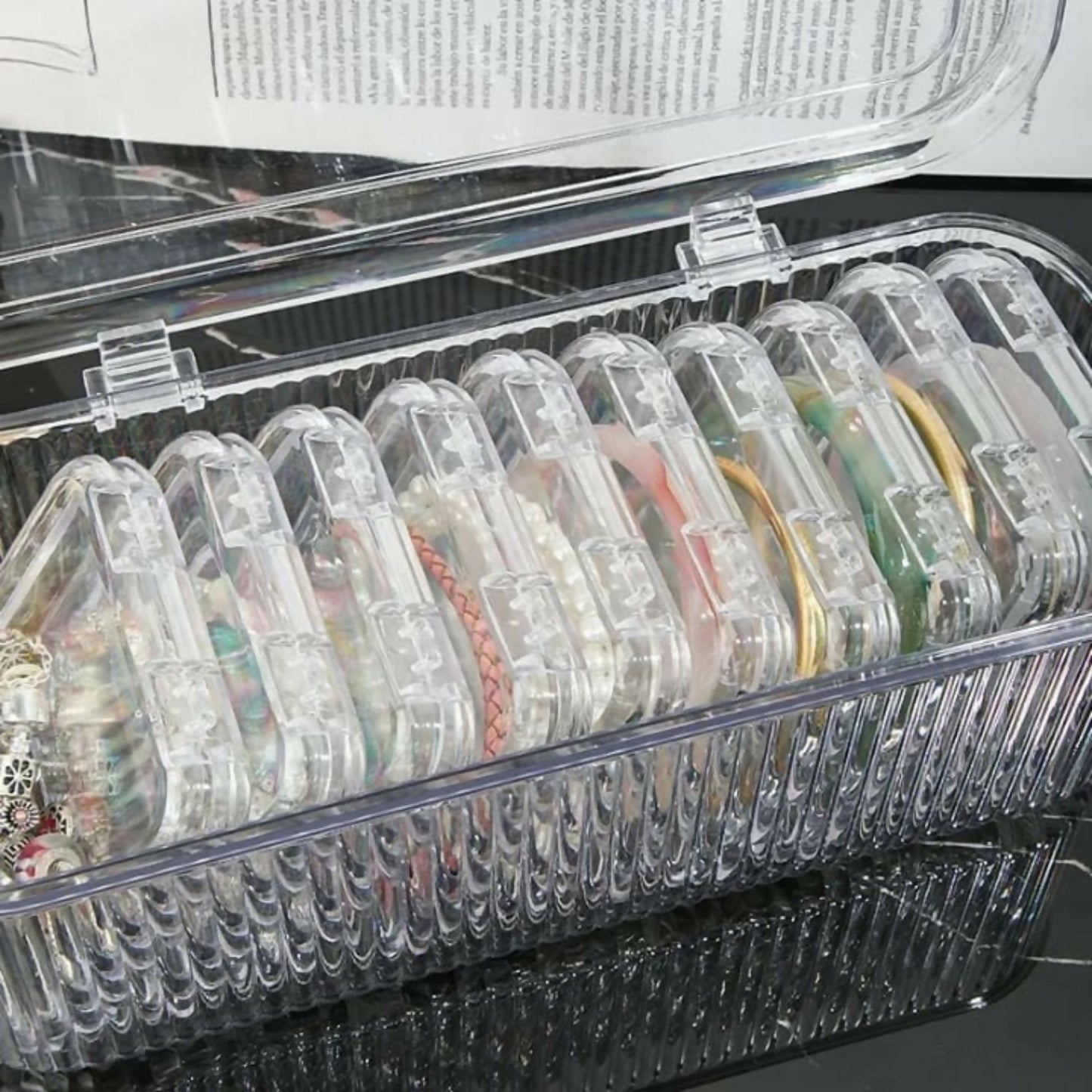 The open clear ribbed storage box reveals multiple small square containers, each holding various pieces of jewelry.