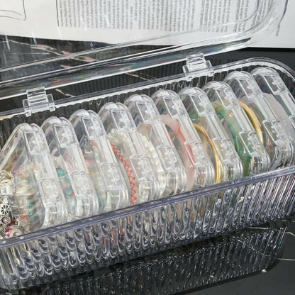 The open clear ribbed storage box reveals multiple small square containers, each holding various pieces of jewelry.
