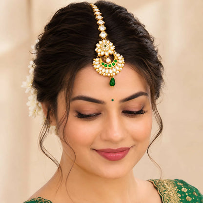 A woman with dark hair models a gold-tone maang tikka with clear stones, green gems, and small white pearls.