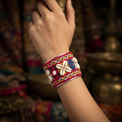 A person's arm wears a wide silver cuff bracelet adorned with red, yellow, and blue embroidery, small mirrors, and white