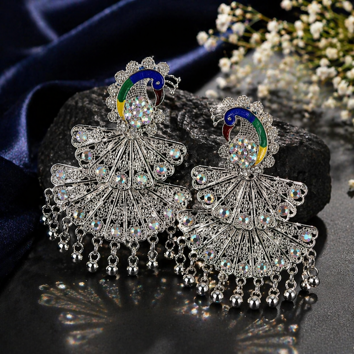 A pair of intricate silver peacock earrings featuring colorful enamel and dangling beads, resting on a dark textured surface.