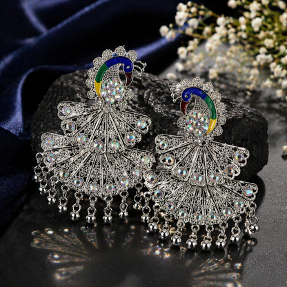 A pair of intricate silver peacock earrings featuring colorful enamel and dangling beads, resting on a dark textured surface.