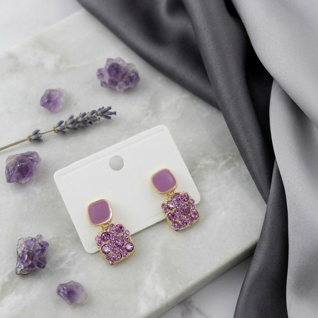 A pair of purple crystal and enamel square drop earrings on a white card, set on a white marble surface with lavender sprigs