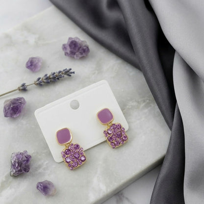 A pair of purple crystal and enamel square drop earrings on a white card, set on a white marble surface with lavender sprigs