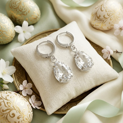 Silver teardrop crystal earrings sit on a small cream pillow, surrounded by decorative eggs and white flowers.