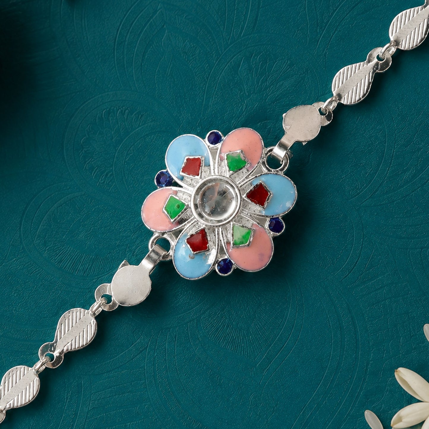 A silver chain rakhi with a colorful enamel flower charm on a dark teal background.