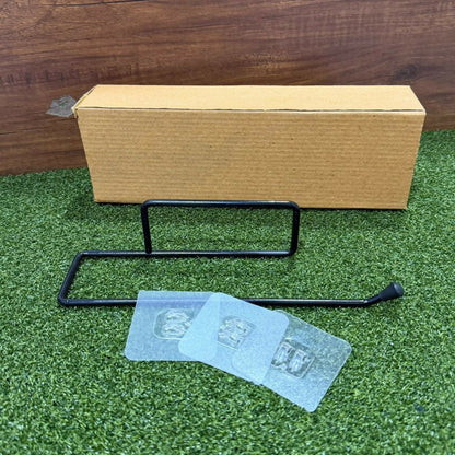 Disassembled black metal paper towel holder with clear adhesive pads and a brown cardboard box on green grass.