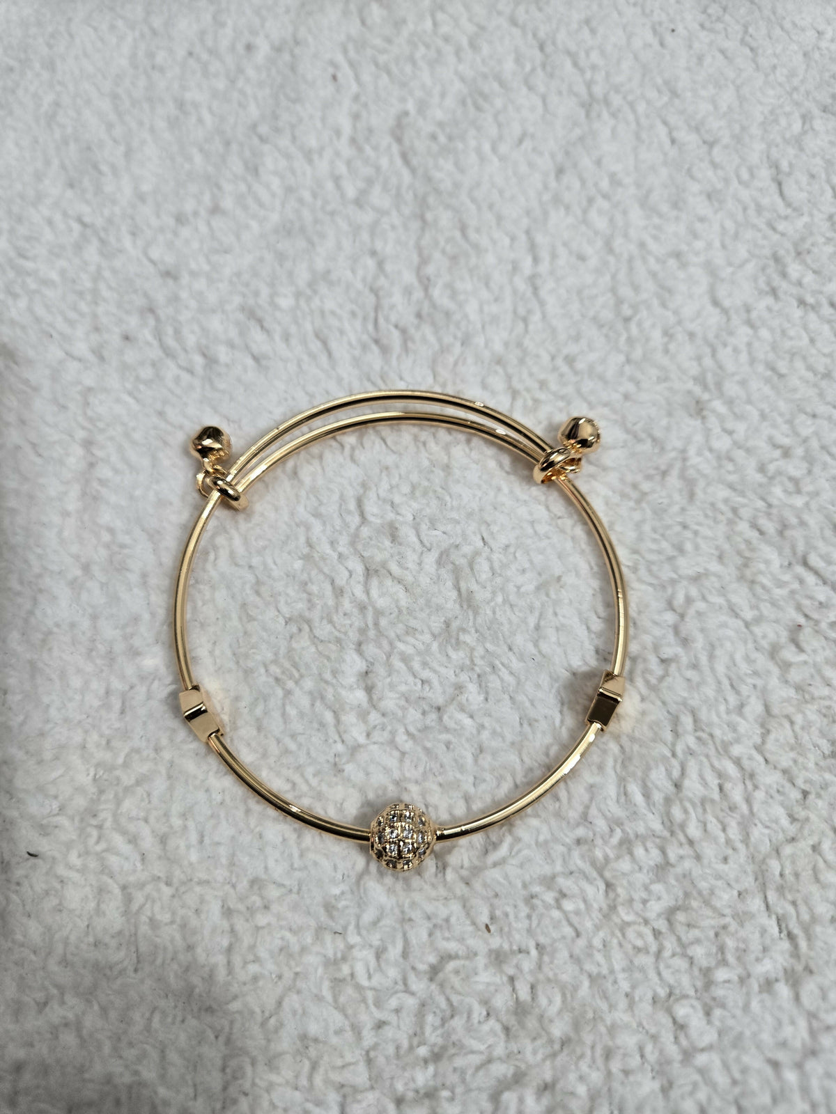 A gold-tone adjustable bangle bracelet with a glittering crystal ball charm lies flat on a textured white fabric.