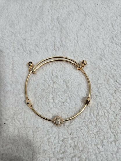 A gold-tone adjustable bangle bracelet with a glittering crystal ball charm lies flat on a textured white fabric.