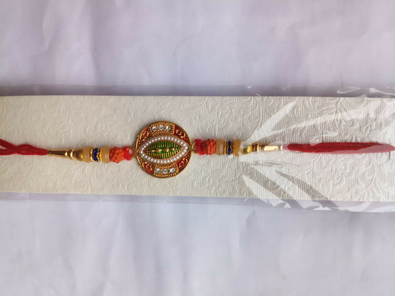 A red and gold bracelet with orange beads, a central green leaf design, and white stones is presented on a textured white