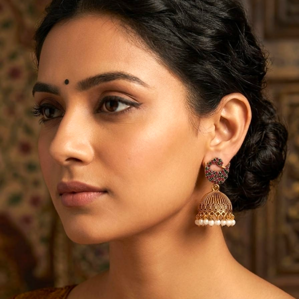 A woman models one gold jhumka earring with red and green stones and pearl drops.