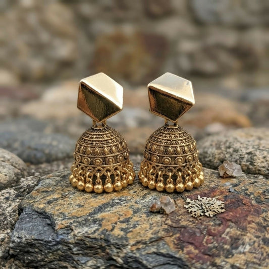A pair of antique gold-plated jhumka earrings with geometric hexagon tops and dome bases, resting on rocks.