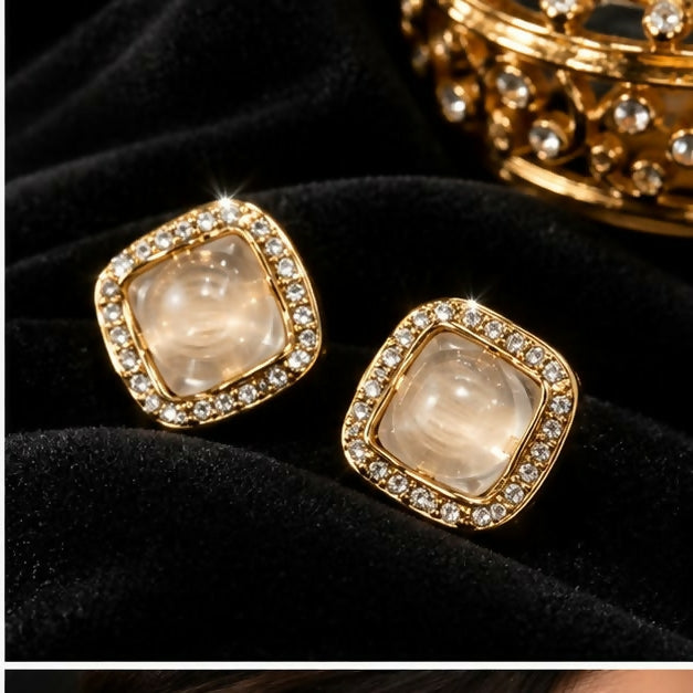 A pair of square stud earrings with clear centers and shimmering gem borders rests on a dark, plush fabric.
