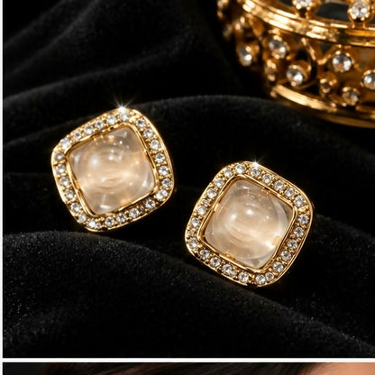A pair of square stud earrings with clear centers and shimmering gem borders rests on a dark, plush fabric.