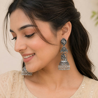 A woman models one of the oxidized silver jhumka earrings, showing the Radha Krishna temple design.