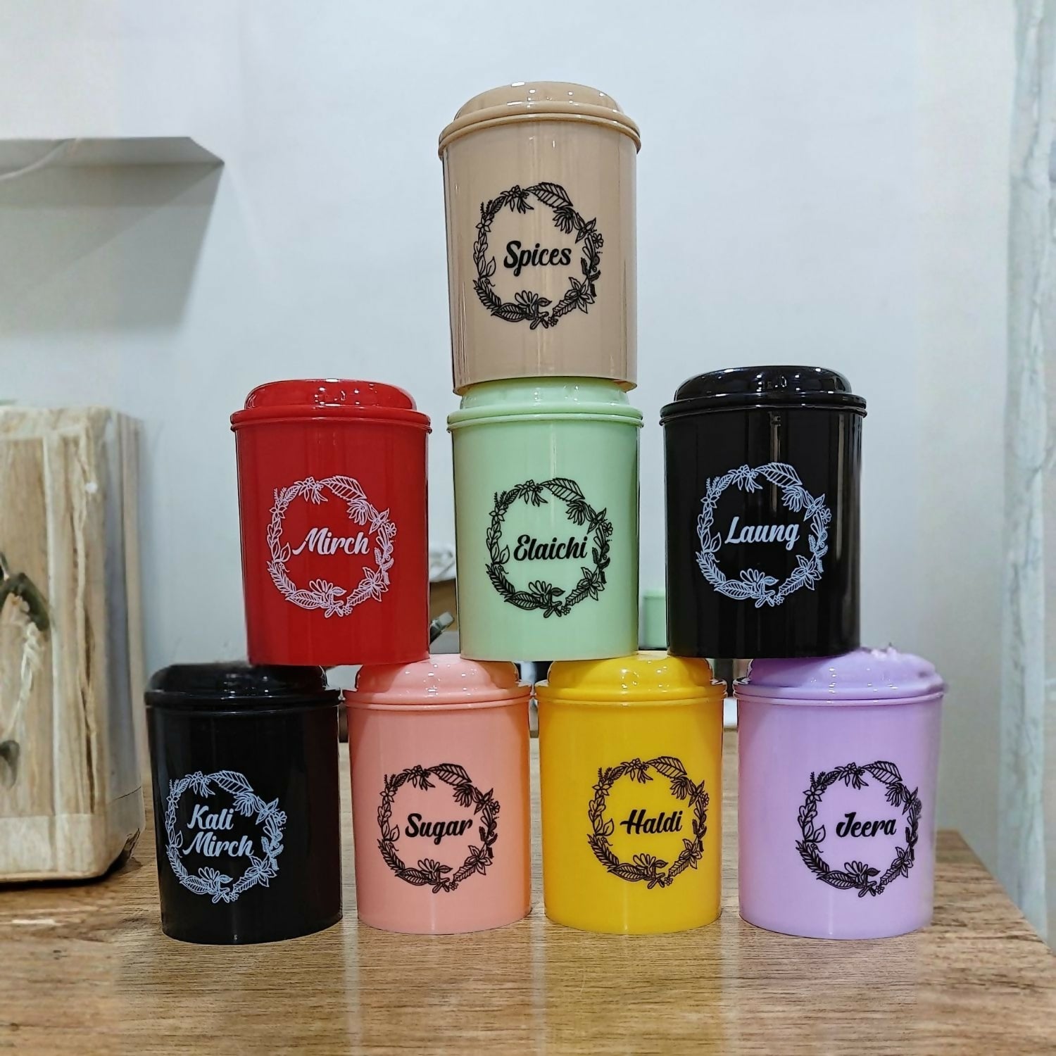 Nine colorful spice containers in black, red, peach, mint, yellow, pink, purple, and gray, arranged in a pyramid on a wooden