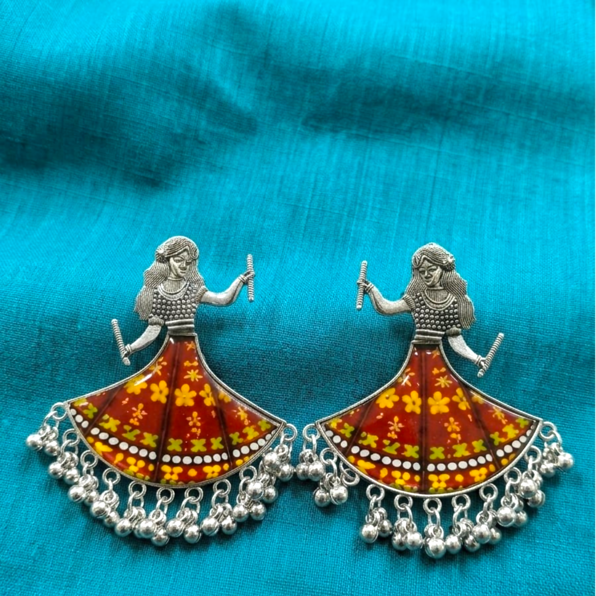 A pair of silver oxidized earrings featuring a dancing girl with a maroon and yellow floral skirt, set on a blue fabric.