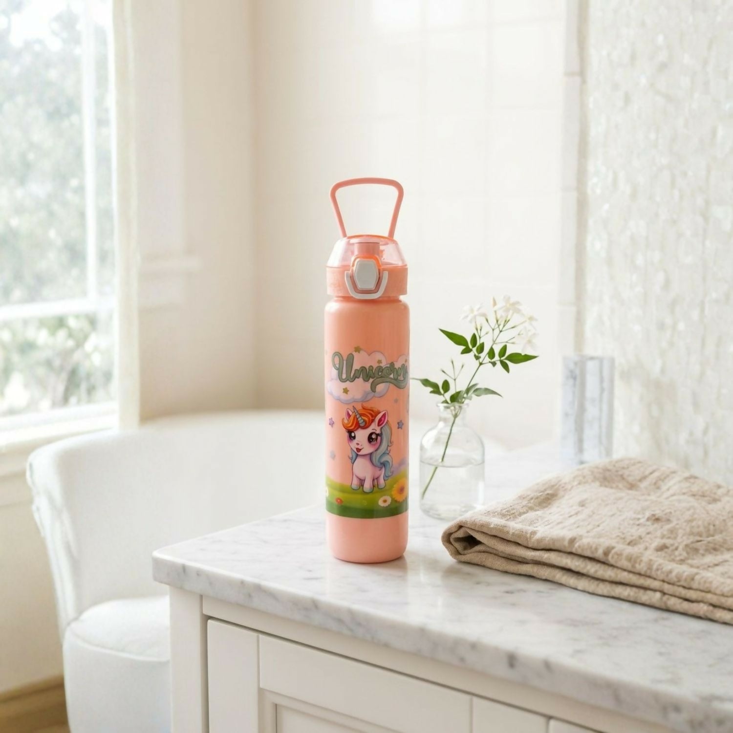 A peach-colored unicorn print water bottle with a straw and carry handle sits on a white marble countertop indoors.