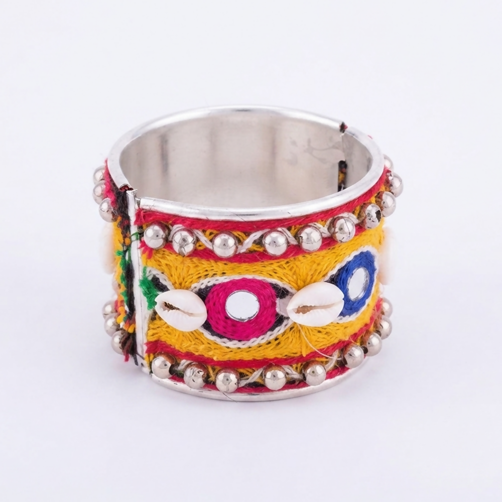 A front view of the wide metal cuff bracelet, featuring intricate thread work, small round mirrors, and white cowrie shells