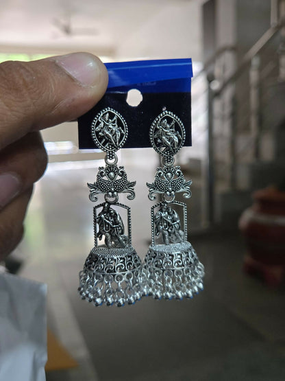 A hand holds up a pair of oxidized silver jhumka earrings with Radha Krishna temple design against a blurry background.