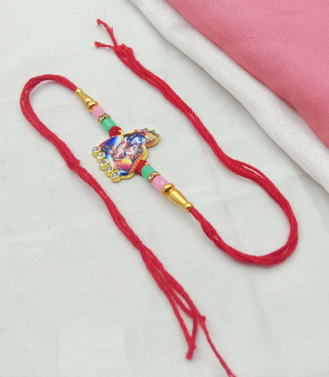 A red thread with a golden charm featuring Radha Krishna, rhinestones, and pastel beads, on a white and pink fabric.