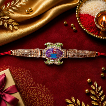 A rose gold Rakhi with a purple, blue, and green Shivling design is centered on a gold cloth with a red border.