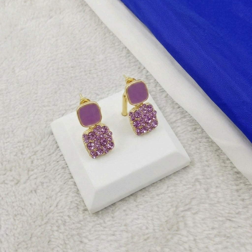 A pair of purple crystal and enamel square drop earrings on a white display stand, with a blue and white fabric in the