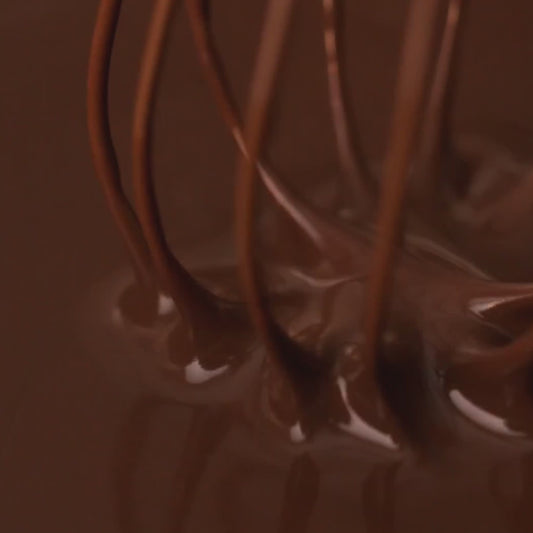 A close-up view of a metal whisk dripping rich, dark chocolate into a pool of melted chocolate.