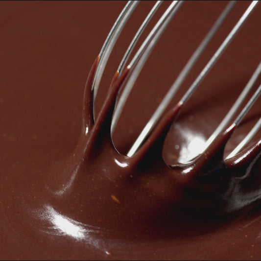 A close-up view of a metal whisk stirring glossy, rich chocolate ganache in a bowl.