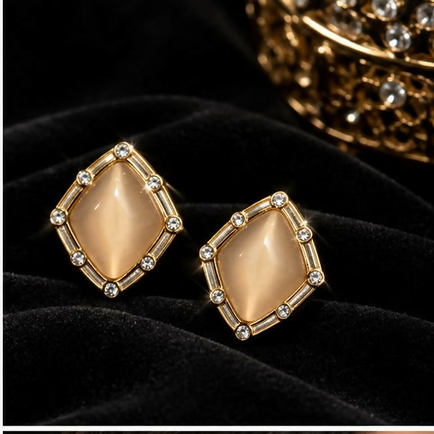 Two champagne geometric stud earrings with gold-tone frames and clear stone accents are displayed on a dark velvet cloth.