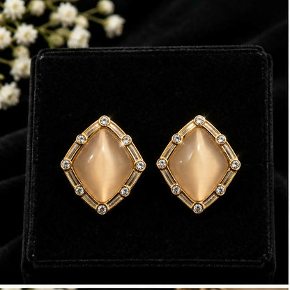A close-up of two champagne geometric stud earrings with gold-tone frames and clear stone accents in a black velvet box.