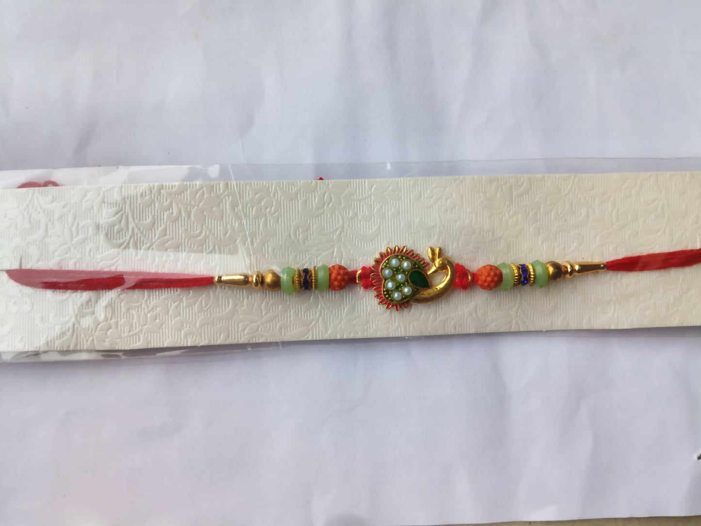 A gold peacock Rakhi with green and pearl details, flanked by orange and mint green beads, on a red thread, displayed on a
