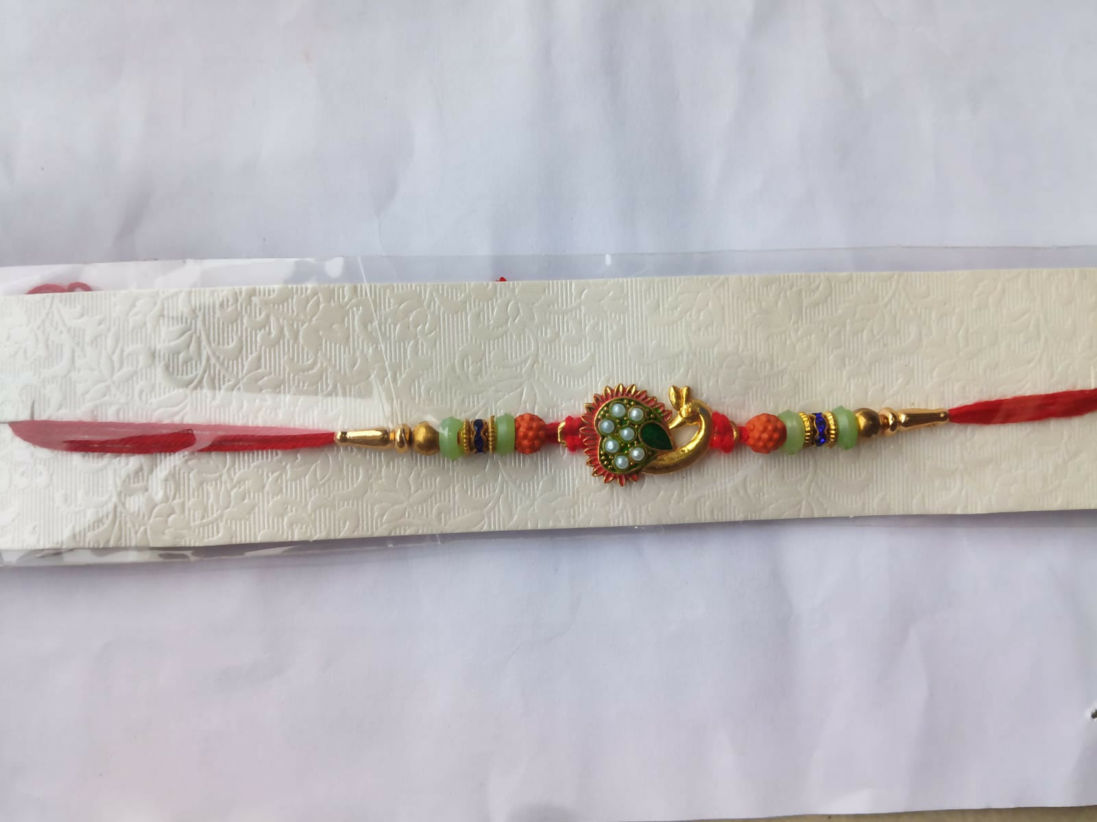 A gold peacock Rakhi with green and pearl details, flanked by orange and mint green beads, on a red thread, displayed on a