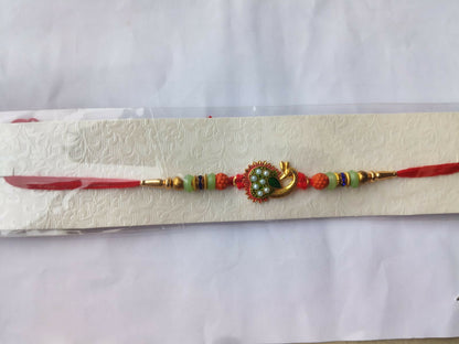 A gold peacock Rakhi with green and pearl details, flanked by orange and mint green beads, on a red thread, displayed on a
