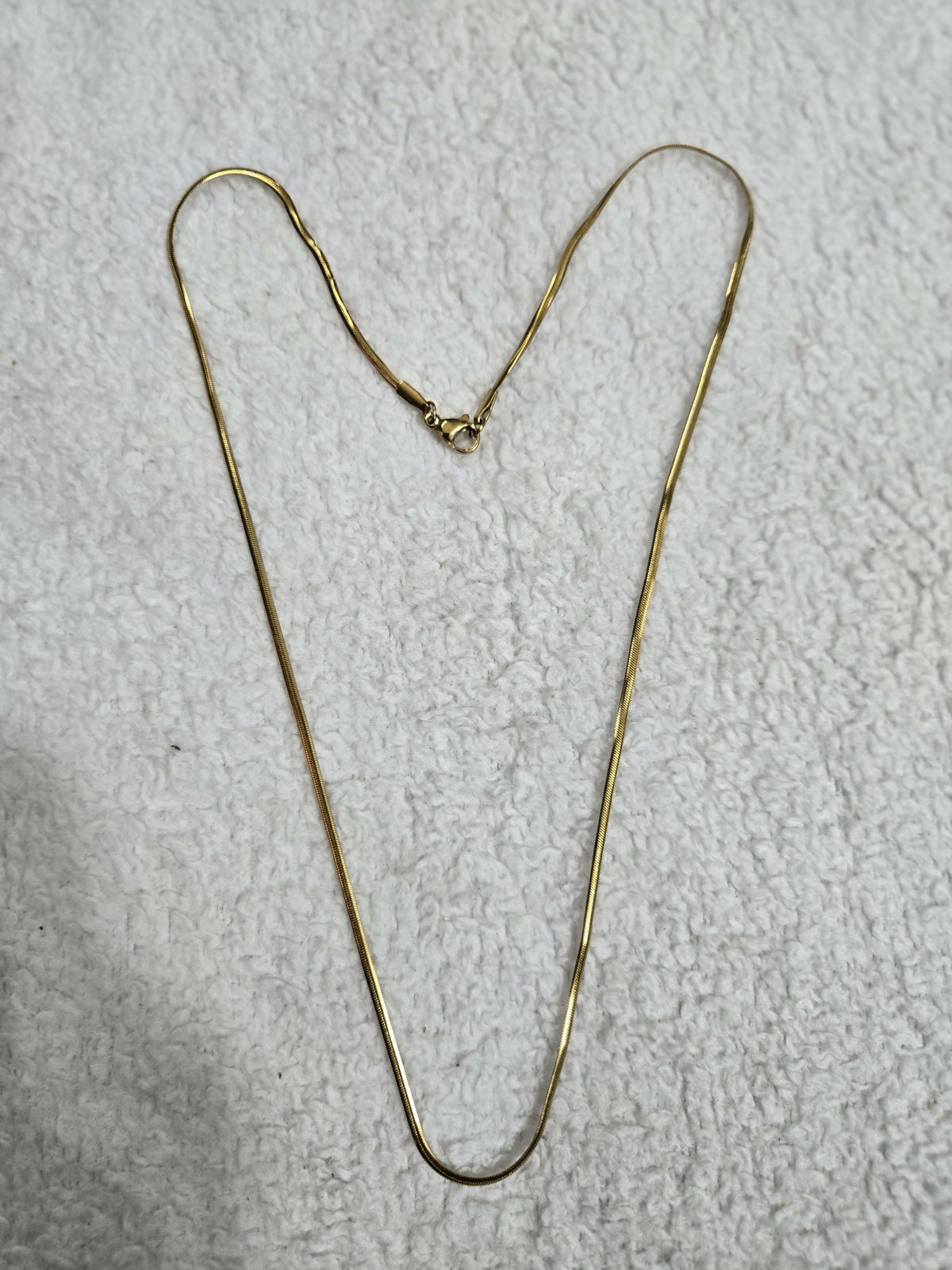 A simple gold-tone snake chain necklace with its clasp is laid out on a textured white cloth surface.