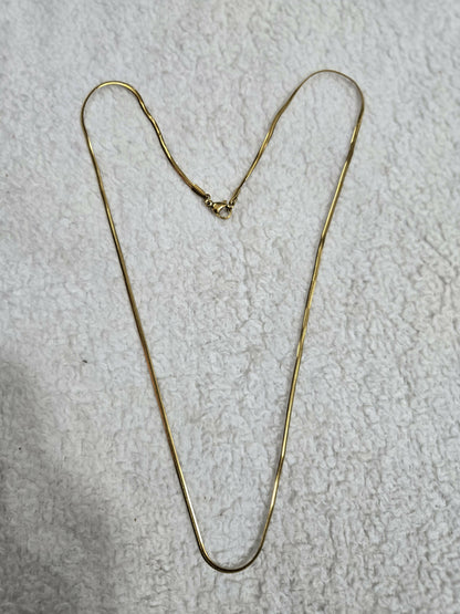 A simple gold-tone snake chain necklace with its clasp is laid out on a textured white cloth surface.